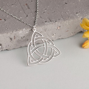 May include: A silver Celtic knot pendant necklace with a triangular shape. The necklace is on a gray textured surface with a yellow flower in the background.