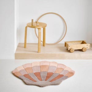 May include: A pink and brown patterned rug shaped like a seashell. The rug is on a light wood floor in a room with a wooden stool and a wooden toy wagon.