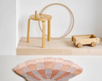 May include: A pink and brown patterned rug shaped like a seashell. The rug is on a light wood floor in a room with a wooden stool and a wooden toy wagon.