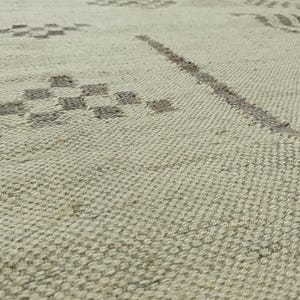 May include: A close-up of a woven rug with a subtle geometric pattern in a light beige color.
