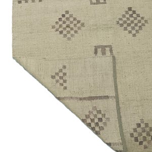 May include: A beige rug with a geometric pattern in brown. The rug has a fringe border and is woven with a textured surface.