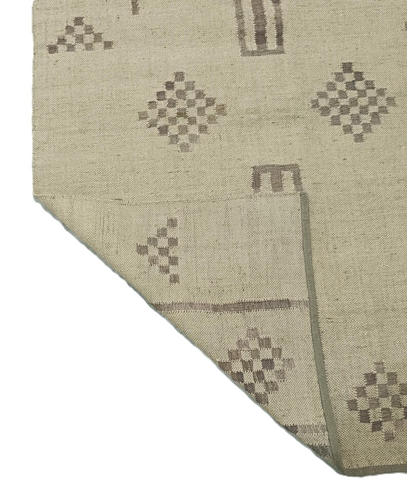 May include: A beige rug with a geometric pattern in brown. The rug has a fringe border and is woven with a textured surface.