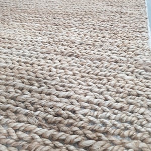 Peut inclure: Un tapis en fibres naturelles fait de corde de jute tissée. Le tapis est de couleur brun clair et a une surface texturée.