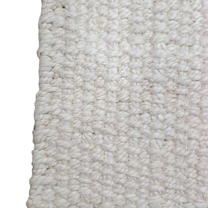 Peut inclure: Gros plan sur un tapis tissé avec une surface texturée blanc cassé. Le tapis présente un tissage serré et noué, avec une bordure bleu foncé le long du bord inférieur. Le design du tapis est simple et neutre, adapté à divers styles d'intérieur.