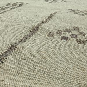 May include: A close-up of a beige woven rug with a geometric pattern in a darker shade of beige. The pattern is made up of lines and squares.