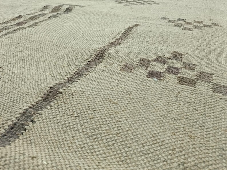 May include: A close-up of a beige woven rug with a geometric pattern in a darker shade of beige. The pattern is made up of lines and squares.