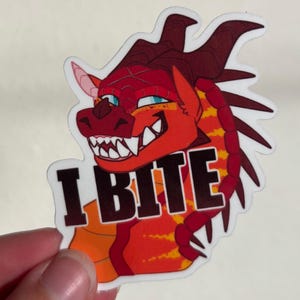 Może przedstawiać: Naklejka przedstawiająca głowę smoka z kreskówki z czerwonymi, pomarańczowymi i żółtymi łuskami. Smok ma ostre białe zęby i niebieskie oczy. Tekst "I BITE" jest wydrukowany pogrubionymi czarnymi literami na białym tle.