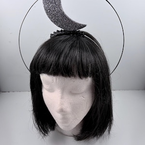 May include: A black headband with a crescent moon and a halo. The moon is covered in silver glitter. The halo is a thin, black wire. The headband is worn on a mannequin head with black hair.