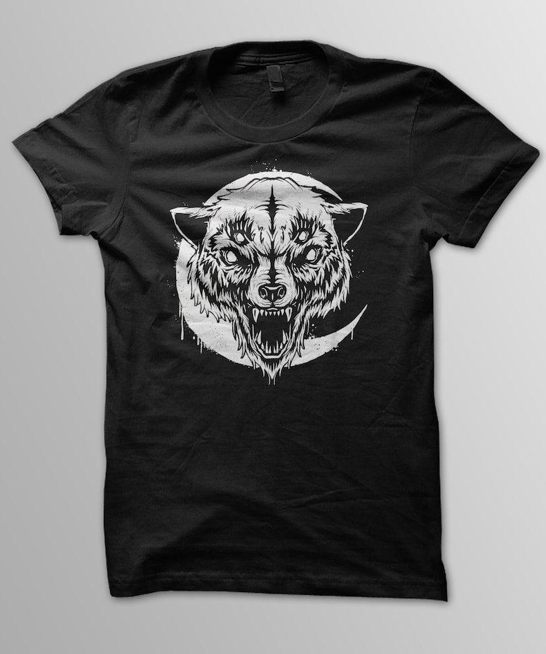 May include: Black t-shirt with a white graphic of a wolf's head with three eyes and a crescent moon behind it.