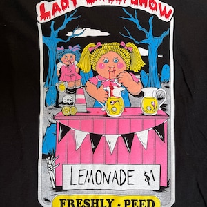 May include: A black t-shirt with a graphic of a doll with red hair and a pink dress selling lemonade. The text on the graphic reads "Lady Creeps Show", "Lemonade $1", and "Freshly - Peed".