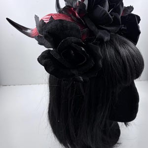May include: A black headpiece featuring black roses, dark gray leaves, and two curved horns with red and pink accents. The headpiece is worn on a black wig. The roses are made of a velvety material.