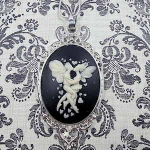 May include: A silver-toned pendant necklace featuring an oval cameo with a black background and a white relief of two cherubs embracing. The pendant is framed with an ornate silver filigree design and hangs from a delicate chain.
