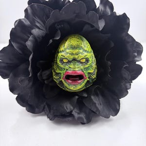 May include: A green and yellow creature face with red lips is centered in a large black flower. The creature has yellow eyes and a textured surface. The flower petals are dark and surround the face.