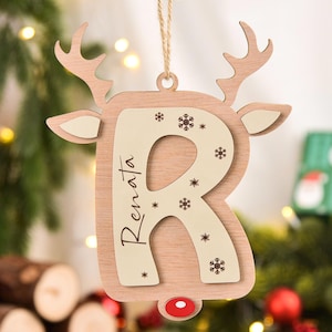 May include: A wooden reindeer Christmas decoration featuring the letter "R" and the name "Renata". The ornament has a light brown base with a cream-coloured centre, snowflake details, and a red nose. It hangs from a natural twine.