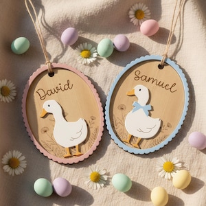 Puede incluir: Dos etiquetas ovaladas de madera con ilustraciones de patos. Una etiqueta tiene un borde rosa y el nombre "David", la otra un borde azul y el nombre "Samuel". Margaritas y huevos de colores pastel rodean las etiquetas.