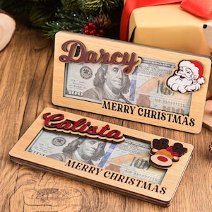 May include: Two rectangular light-colored wooden money holders with cutouts displaying 100-dollar bills. One features the name Darcy and a Santa Claus icon, the other Calista and a reindeer icon. Both are engraved with the text MERRY CHRISTMAS.