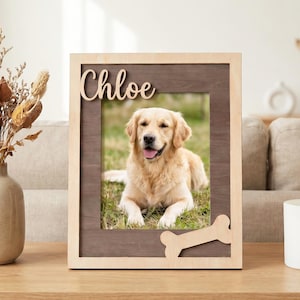 May include: A rectangular wooden picture frame with a light wood outer border and a dark wood inner panel. The frame features the name Chloe in script lettering at the top and a bone-shaped cutout at the bottom right corner, holding a photo of a dog.