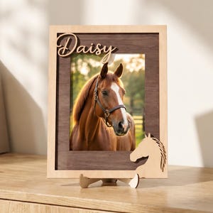 May include: A rectangular wooden picture frame featuring a dark brown inner border and a light wood outer frame. The name Daisy is cut out in script font at the top, and a horse head silhouette is attached to the bottom right corner.