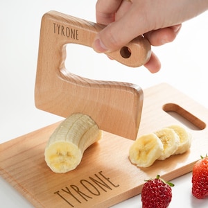 Può includere: Un tagliere di legno con il nome "Tyrone" inciso su di esso. Una banana viene tagliata con un affettatrice di banana in legno, anch'essa incisa con il nome "Tyrone". Ci sono banane a fette e fragole sul tagliere.