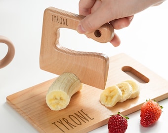 May include: A light-colored wooden kitchen tool with the name TYRONE engraved on the handle, used to slice a banana on a matching wooden cutting board that also features the name TYRONE, accompanied by two fresh red strawberries.