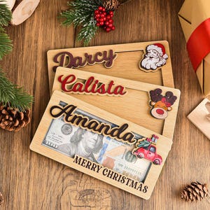 K&ouml;nnte beinhalten: Drei rechteckige Geldgeschenkhalter aus Holz mit Aussparungen f&uuml;r Geldscheine. Jeder ist mit einem personalisierten Namen in Schreibschrift versehen, darunter Darcy, Calista und Amanda, sowie festlichen Motiven wie Weihnachtsmann und Rentier.