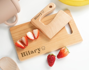 May include: A rectangular wooden cutting board and a matching wooden toy knife, both engraved with the name Hilary and a chef hat icon. The board holds sliced strawberries, with additional whole strawberries placed nearby on the surface.
