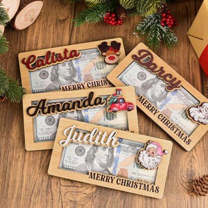 K&ouml;nnte beinhalten: Vier rechteckige Geldgeschenkhalter aus Holz mit Aussparungen f&uuml;r Hundert-Dollar-Scheine. Jeder Halter zeigt einen personalisierten Namen in Schreibschrift, den Text MERRY CHRISTMAS und ein festliches Holzmotiv wie Rentier, LKW oder Weihnachtsmann.
