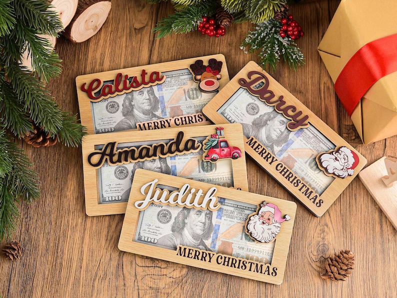 K&ouml;nnte beinhalten: Vier rechteckige Geldgeschenkhalter aus Holz mit Aussparungen f&uuml;r Hundert-Dollar-Scheine. Jeder Halter zeigt einen personalisierten Namen in Schreibschrift, den Text MERRY CHRISTMAS und ein festliches Holzmotiv wie Rentier, LKW oder Weihnachtsmann.