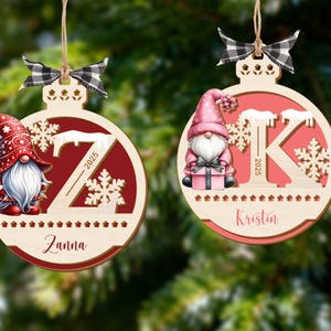May include: Two wooden Christmas tree decorations with gnome designs. One features a red gnome with a white beard and a red hat, the other a pink gnome. Both have a letter, snowflakes, and the year 2025. Each decoration has a name at the bottom.