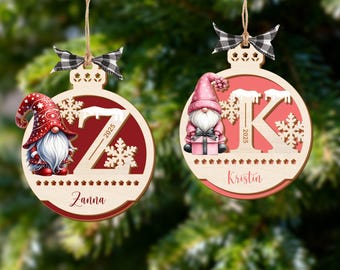 Könnte beinhalten: Zwei Weihnachtsbaumschmuck aus Holz mit Zwergenmotiven. Einer zeigt einen roten Zwerg mit weißem Bart und roter Mütze, der andere einen rosa Zwerg. Beide haben einen Buchstaben, Schneeflocken und das Jahr 2025. Jeder Schmuck hat unten einen Namen.