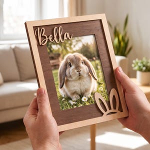 May include: A rectangular wooden picture frame featuring a dark brown wood grain center panel and a light wood border. The name Bella is cut out in script lettering at the top left, and a pair of rabbit ears is cut out at the bottom right corner.