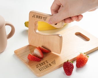 May include: A light-colored wooden pizza cutter with a handle engraved with the name JENNY and a pizza slice icon. The wooden cutting board features the text JENNY'S KITCHEN and a pizza slice icon, with sliced strawberries on the surface.