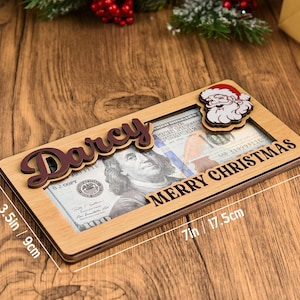 K&ouml;nnte beinhalten: Ein rechteckiger Geldhalter aus Holz mit dem Namen Darcy in dunkelbrauner Schreibschrift, einem Weihnachtsmann-Ausschnitt und dem Text Merry Christmas. Er misst 9 cm mal 17,5 cm und enth&auml;lt einen 100-Dollar-Schein im mittigen Ausschnitt.