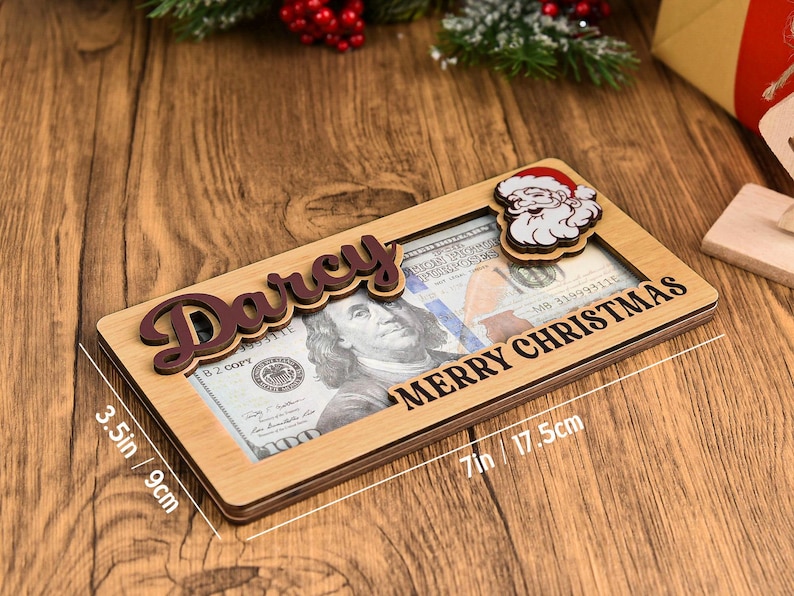 K&ouml;nnte beinhalten: Ein rechteckiger Geldhalter aus Holz mit dem Namen Darcy in dunkelbrauner Schreibschrift, einem Weihnachtsmann-Ausschnitt und dem Text Merry Christmas. Er misst 9 cm mal 17,5 cm und enth&auml;lt einen 100-Dollar-Schein im mittigen Ausschnitt.