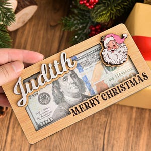 K&ouml;nnte beinhalten: Ein rechteckiger Geldhalter aus hellem Holz mit einem Ausschnitt, in dem ein Hundert-Dollar-Schein zu sehen ist. Der Halter tr&auml;gt den Namen Judith in wei&szlig;er Schreibschrift und den Text MERRY CHRISTMAS in Schwarz sowie ein kleines Weihnachtsmann-Symbol.