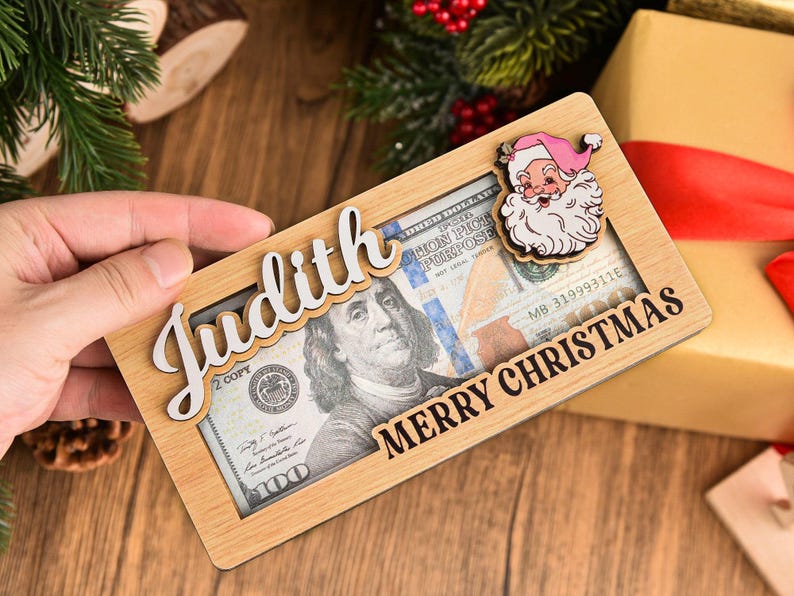 K&ouml;nnte beinhalten: Ein rechteckiger Geldhalter aus hellem Holz mit einem Ausschnitt, in dem ein Hundert-Dollar-Schein zu sehen ist. Der Halter tr&auml;gt den Namen Judith in wei&szlig;er Schreibschrift und den Text MERRY CHRISTMAS in Schwarz sowie ein kleines Weihnachtsmann-Symbol.