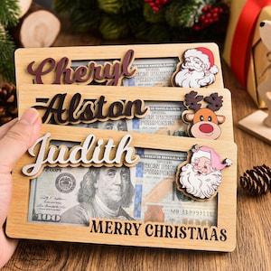 K&ouml;nnte beinhalten: Drei rechteckige Geldhalter aus hellem Holz mit Aussparungen f&uuml;r Geldscheine. Jeder zeigt einen Namen in erhabener Schreibschrift: Cheryl, Alston und Judith. Dekorative Symbole sind der Weihnachtsmann und ein Rentier. Der Text Merry Christmas ist aufgedruckt.