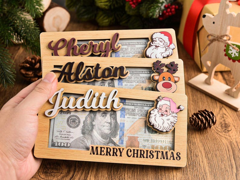 K&ouml;nnte beinhalten: Drei rechteckige Geldhalter aus hellem Holz mit Aussparungen f&uuml;r Geldscheine. Jeder zeigt einen Namen in erhabener Schreibschrift: Cheryl, Alston und Judith. Dekorative Symbole sind der Weihnachtsmann und ein Rentier. Der Text Merry Christmas ist aufgedruckt.