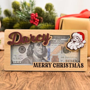 K&ouml;nnte beinhalten: Ein rechteckiger Geldgeschenkrahmen aus hellem Holz mit dem Namen Darcy in dunkler Schreibschrift oben und dem Text MERRY CHRISTMAS in Schwarz unten. Eine Weihnachtsmann-Figur ist an der rechten Seite des Rahmens befestigt.