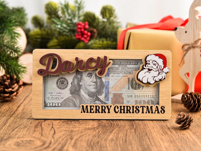 K&ouml;nnte beinhalten: Ein rechteckiger Geldgeschenkrahmen aus hellem Holz mit dem Namen Darcy in dunkler Schreibschrift oben und dem Text MERRY CHRISTMAS in Schwarz unten. Eine Weihnachtsmann-Figur ist an der rechten Seite des Rahmens befestigt.