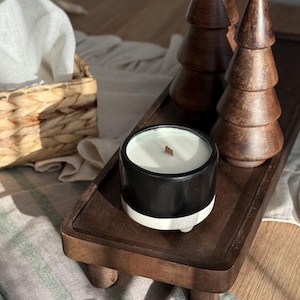 May include: A black and white candle with a wooden wick sits on a dark brown wooden tray, alongside two wooden Christmas tree decorations. The tray rests on a striped fabric, creating a cosy, seasonal display.