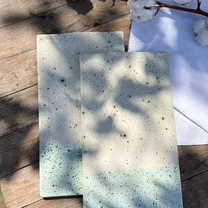 Könnte beinhalten: Zwei rechteckige Keramikuntersetzer mit einem gesprenkelten, zweifarbigen Design. Der obere Teil ist cremefarben und geht in ein gesprenkeltes Türkis am unteren Rand über. Die Untersetzer werden auf einer Holzoberfläche mit Baumwollzweigen im Hintergrund präsentiert.