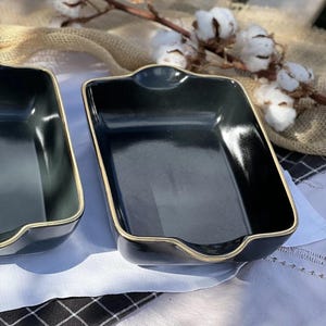 May include: Two rectangular, dark grey ceramic dishes with a gold trim. The dishes have a slightly curved design and are placed on a white tablecloth. Cotton branch in the background.