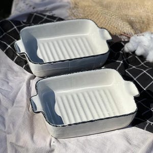 Puede incluir: Dos fuentes para hornear de cerámica blanca con bordes negros y fondo acanalado. Las fuentes están sobre una tela a cuadros blancos y negros.