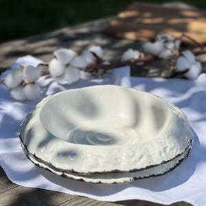 May include: A white ceramic bowl with a textured, organic-shaped rim. The bowl is slightly indented in the center and has a black rim.