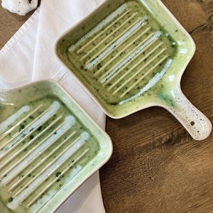May include: Two square ceramic baking dishes with green and white glaze. The dishes have a raised grill pattern on the bottom.