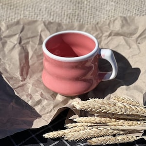Pode incluir: Uma caneca de cerâmica rosa com uma borda branca e uma alça branca. A caneca tem um design texturizado e ondulado e está sobre uma superfície de papel marrom.