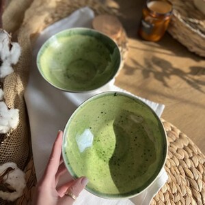Op de afbeelding: Twee groene keramische kommen met een gespikkelde afwerking. De kommen hebben een glanzend glazuur en staan op een witte doek en een geweven placemat. De kommen zijn lichtgroen met donkergroene spikkels.