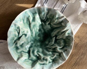 May include: A handcrafted ceramic bowl with a distinctive, textured pattern. The bowl showcases a mix of teal and green shades, with a glossy surface. The edge has a slightly uneven form, enhancing its artisanal appeal. The bowl rests on a white and blue striped cloth.