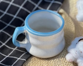 Puede incluir: Una taza de cerámica blanca con un borde azul claro. La taza tiene una superficie texturizada y está sentada sobre una superficie de arpillera.